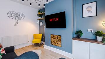 A modern living room with a sofa, a yellow chair, a TV mounted on the wall, a geometric wall art piece, and a wooden cabinet with plants.