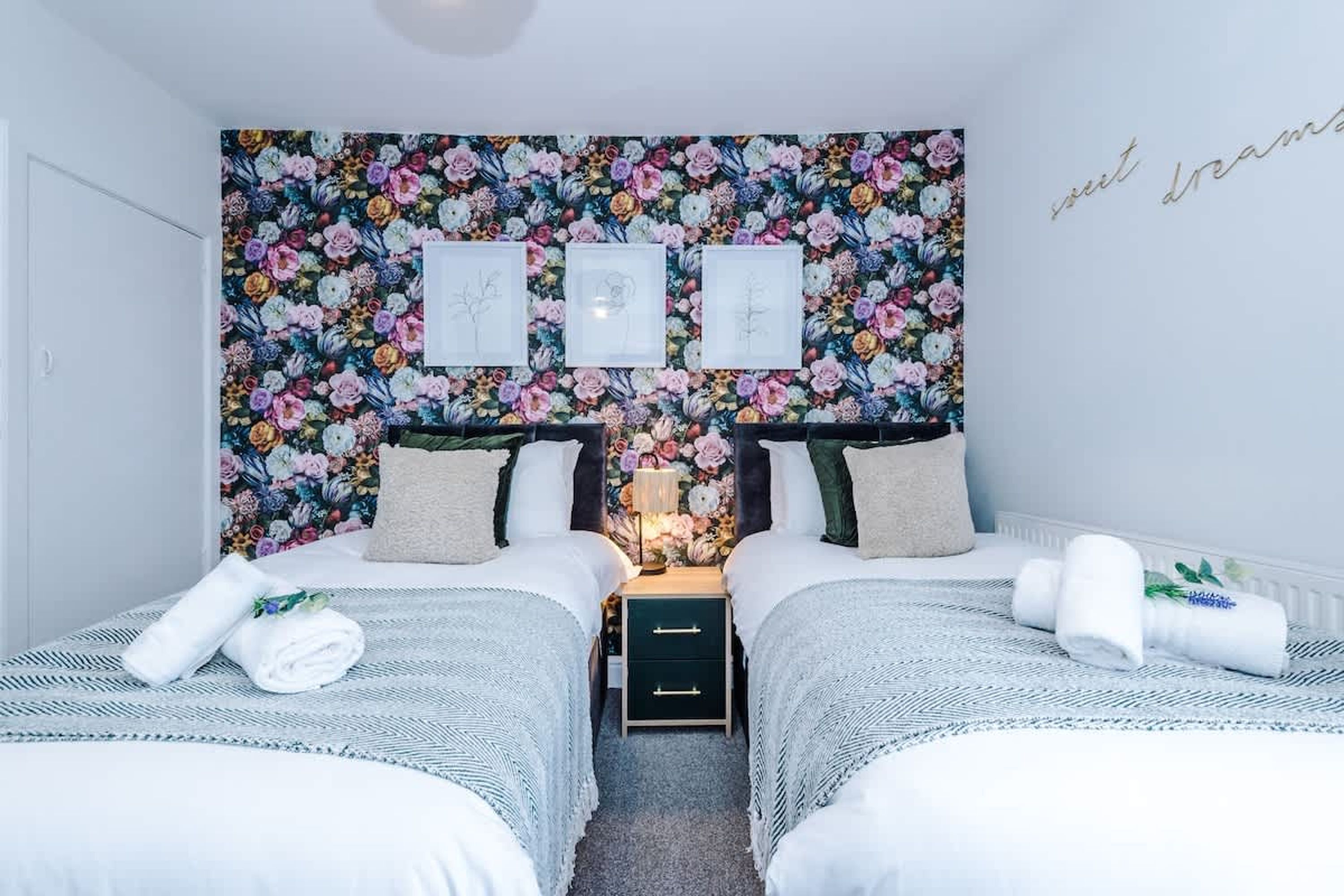 A room with two beds, a floral wallpaper, framed artwork, and a bedside table with a lamp.