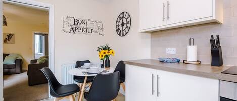 A modern kitchen with white cabinets, a dining area with black chairs, a clock, and a wall art piece.