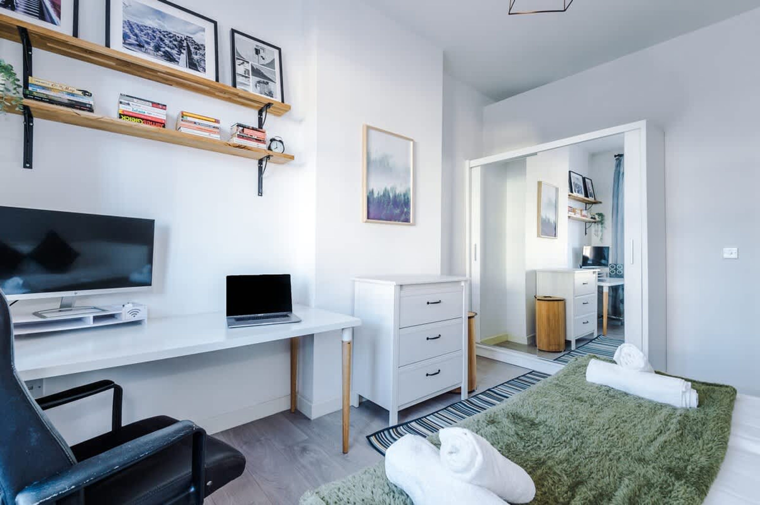A modern hotel room with a flat-screen TV, a laptop, a desk, a white dresser, a green rug, and a bed with towels.