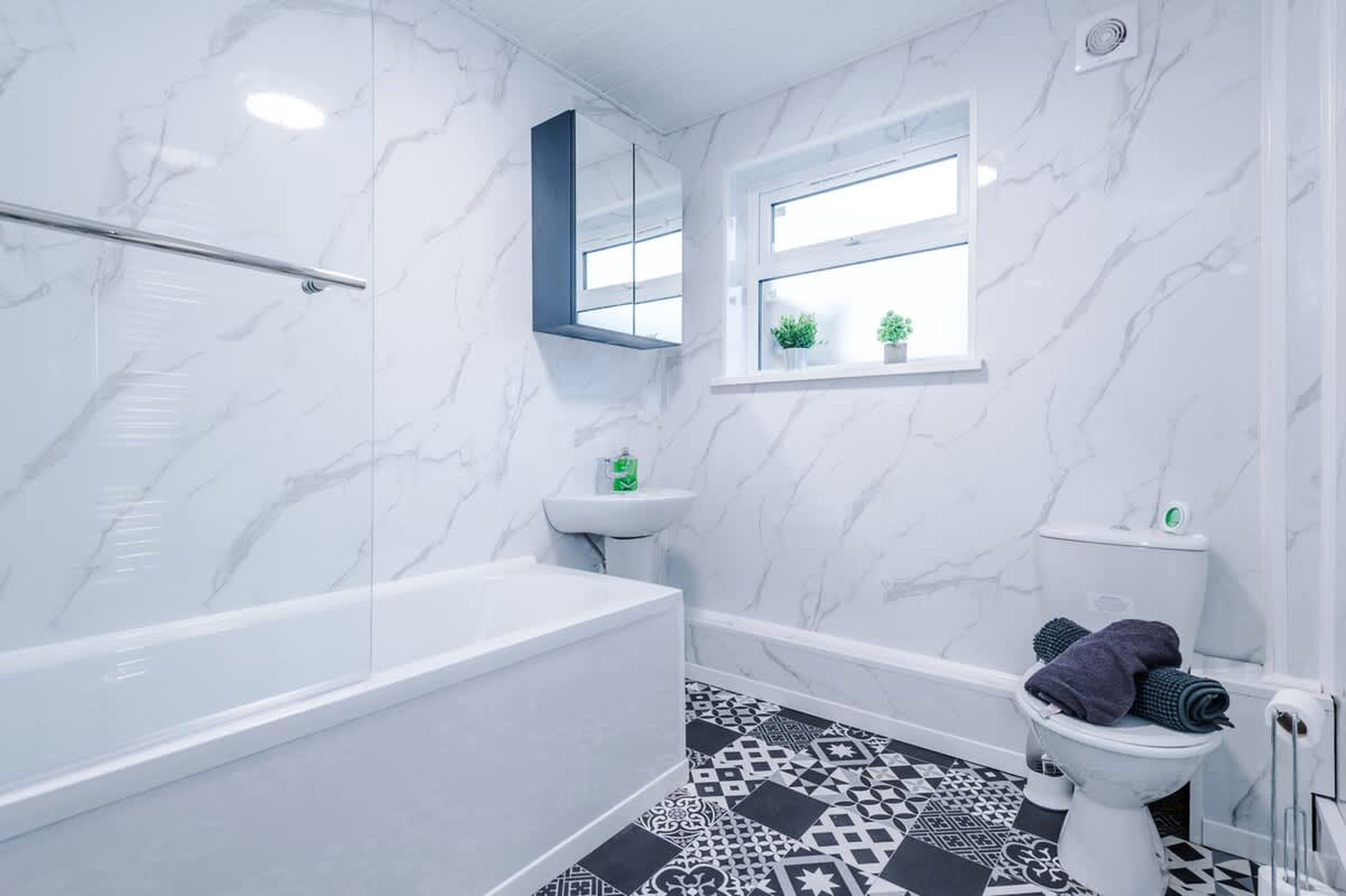 A bathroom with a marble wall, a bathtub, a toilet, a sink, and a window with plants.