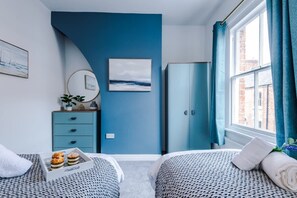 A room with two beds, a blue wall, a wardrobe, a window with curtains, and a tray with pastries. - NEW Stylish City Centre Home Great Location (Chester)