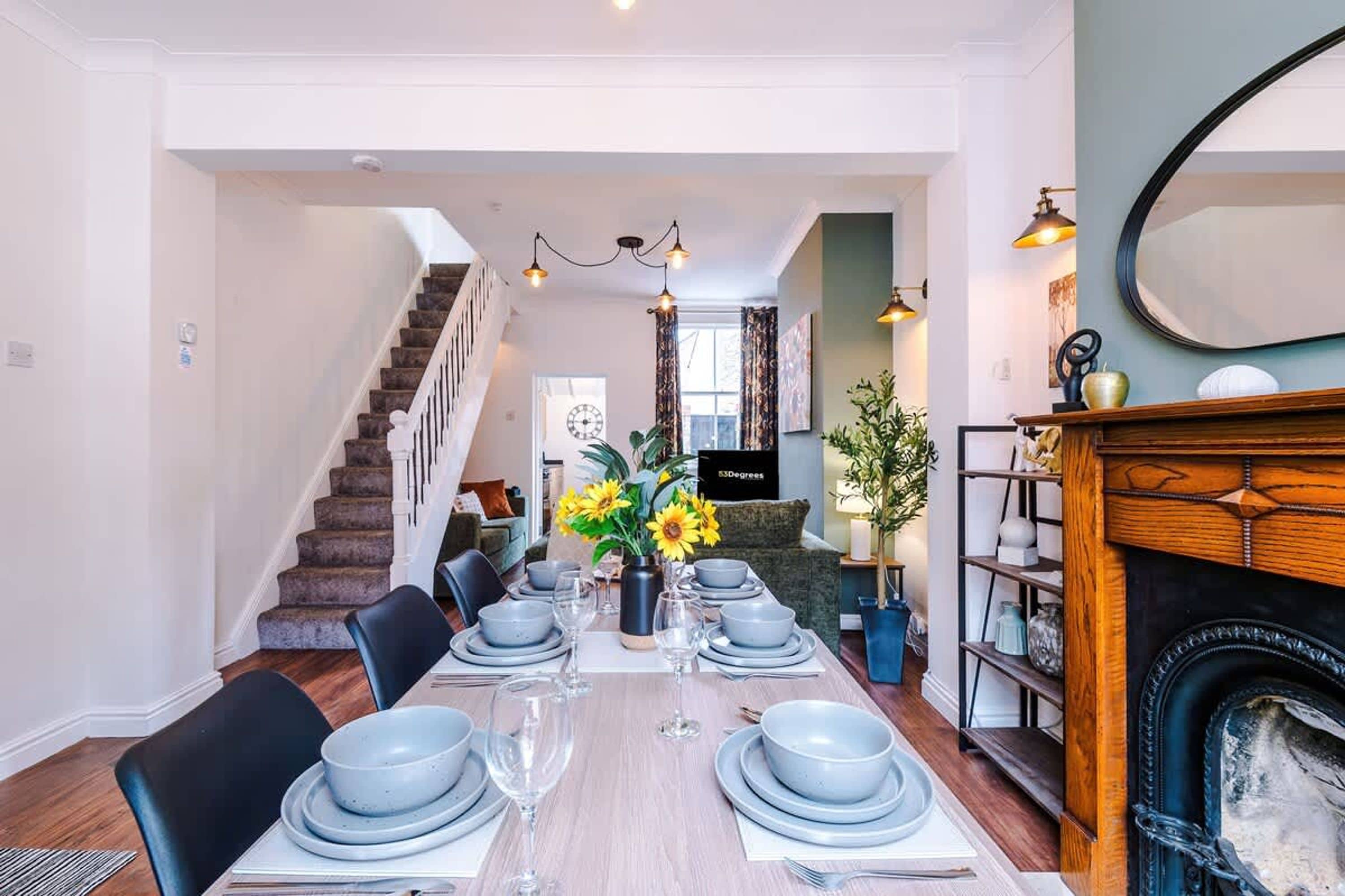 A dining room with a long table set for six, a fireplace, and a staircase leading to another room.