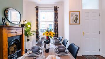 A dining room with a wooden table set for six, a fireplace, a large mirror, and a window with curtains.