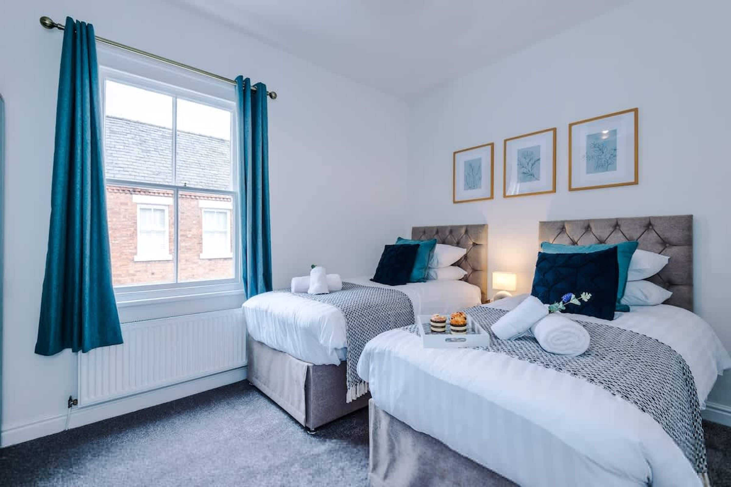 A hotel room with two beds, a window with curtains, a radiator, and three framed pictures on the wall.