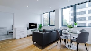 A modern living room with a sofa, dining table, chairs, a TV, and large windows. - NEW Stylish Apartment Amazing Location Sleeps 4 (Manchester)
