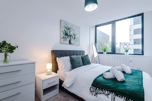 A modern hotel room with a bed, bedside table, lamp, and a window with a view of buildings. - NEW Stylish Apartment Amazing Location Sleeps 4 (Manchester)