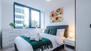 A modern bedroom with a large bed, a window with a view, a painting of sunflowers, and a bedside table with a lamp. - NEW Stylish Apartment Amazing Location Sleeps 4 (Manchester)