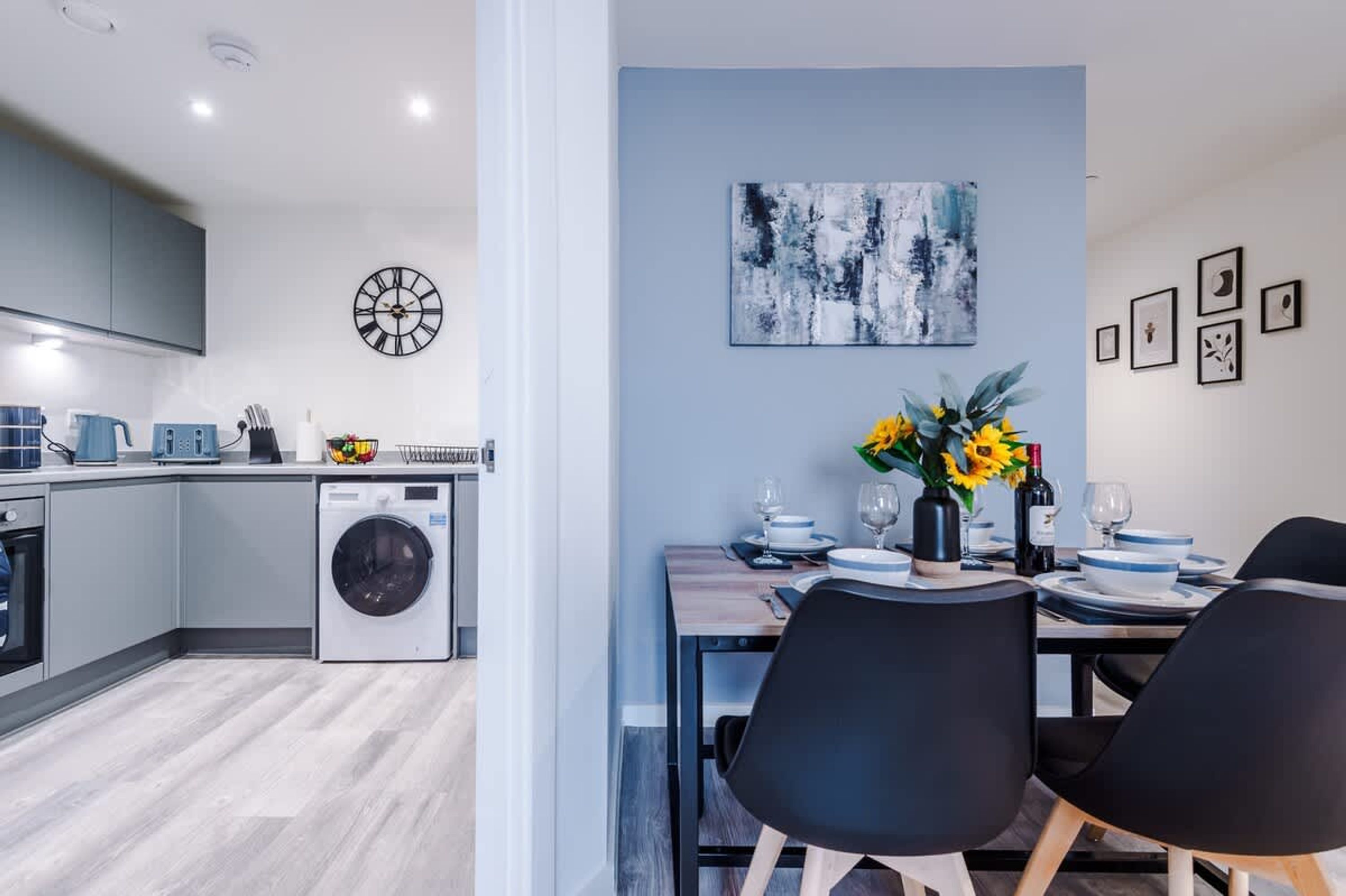 A modern kitchen with a washing machine, a dining area with a table set for two, and a wall with abstract art and framed pictures.