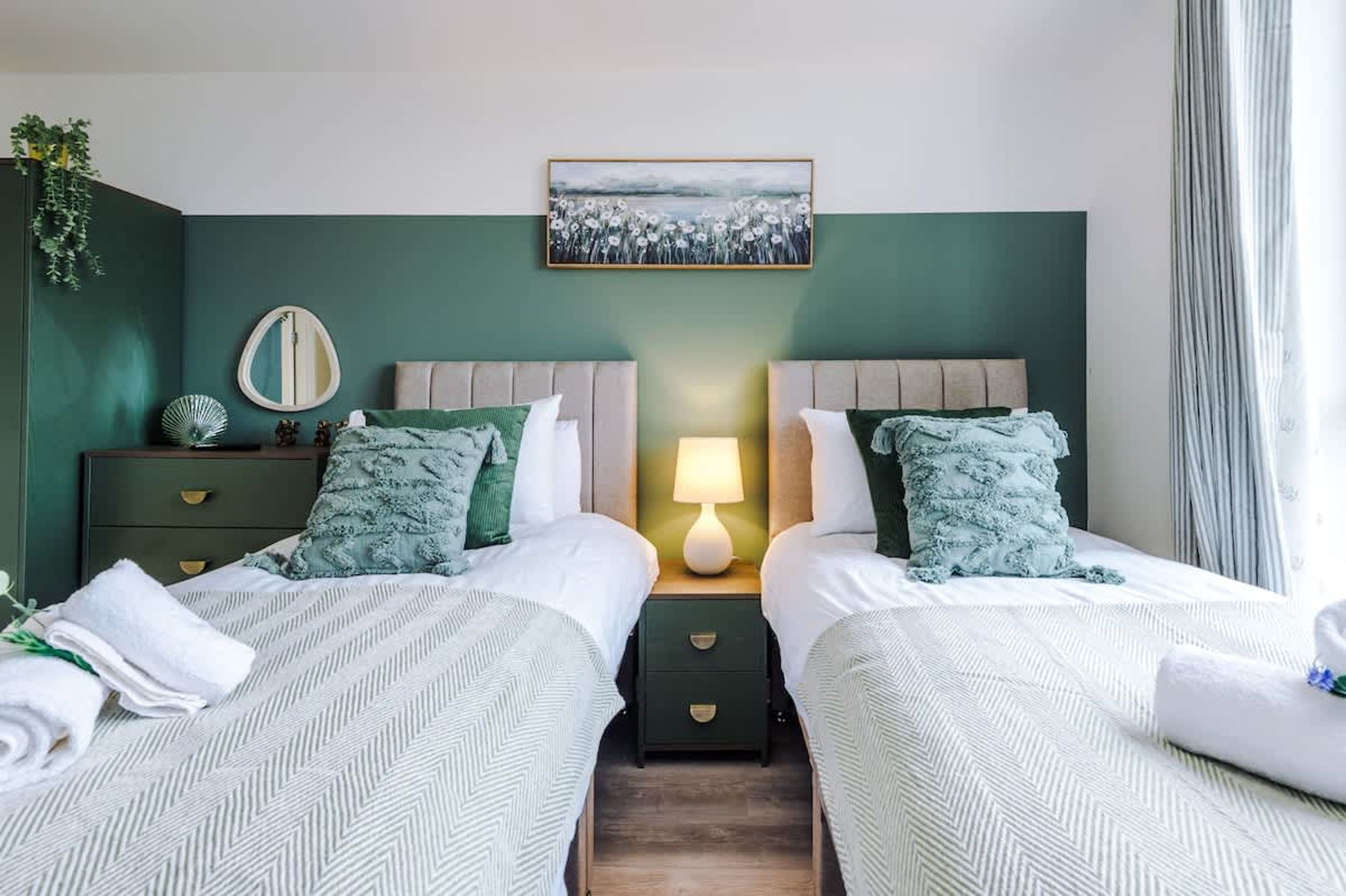A twin room with two beds, a bedside table with a lamp, a mirror, and a painting on the wall.
