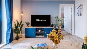 A living room with a flat-screen TV, a blue cabinet, a plant, a vase with dried flowers, and a bottle of champagne. - NEW Pristine 2-bed Flat Great Location Sleeps 6 (Liverpool)