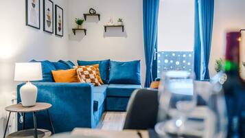 Un salon moderne avec un canapé bleu, une petite table ronde surmontée d’une lampe et une fenêtre ornée de rideaux bleus.