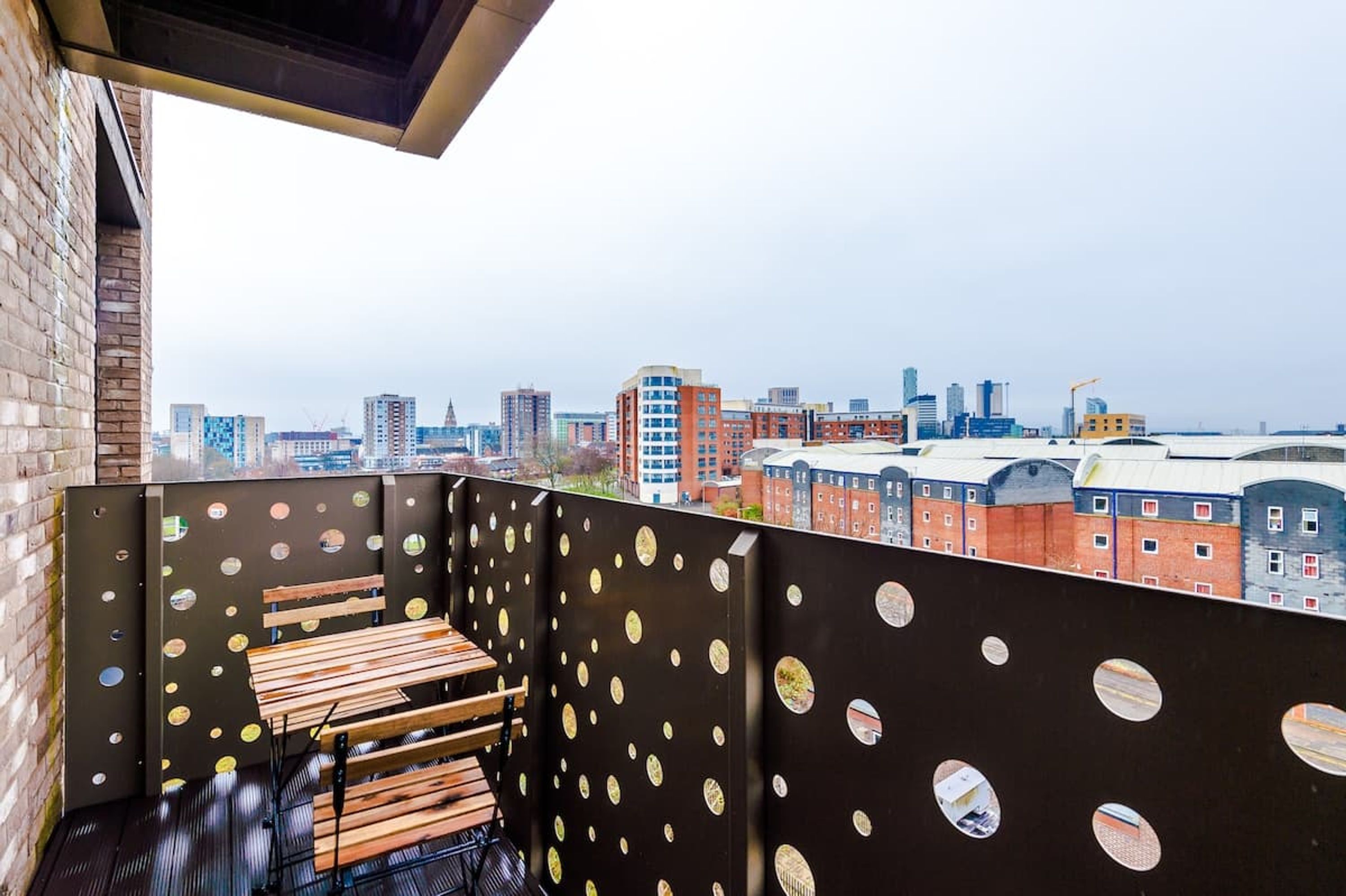 A balcony with a city view, a wooden bench, and a metal wall with circular cutouts.