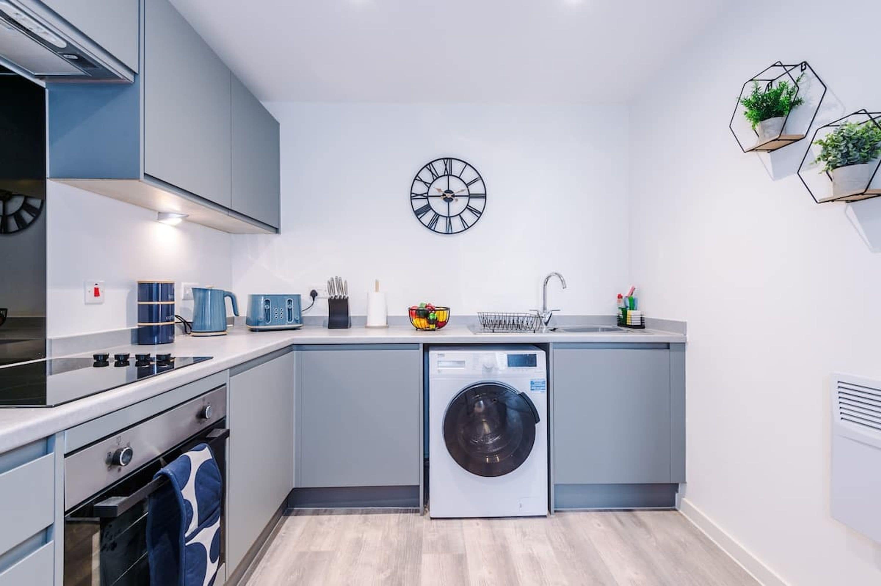 A compact kitchen with a built-in washing machine, a clock on the wall, and a small plant shelf.