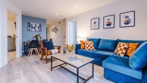 A modern living room with a blue sofa, a coffee table, and a dining area with chairs and a table. - NEW Pristine 2-bed Flat Great Location Sleeps 6 (Liverpool)