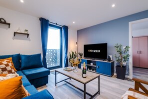 A modern living room with a blue sofa, a flat-screen TV, a coffee table, and a balcony with blue curtains. - NEW Pristine 2-bed Flat Great Location Sleeps 6 (Liverpool)