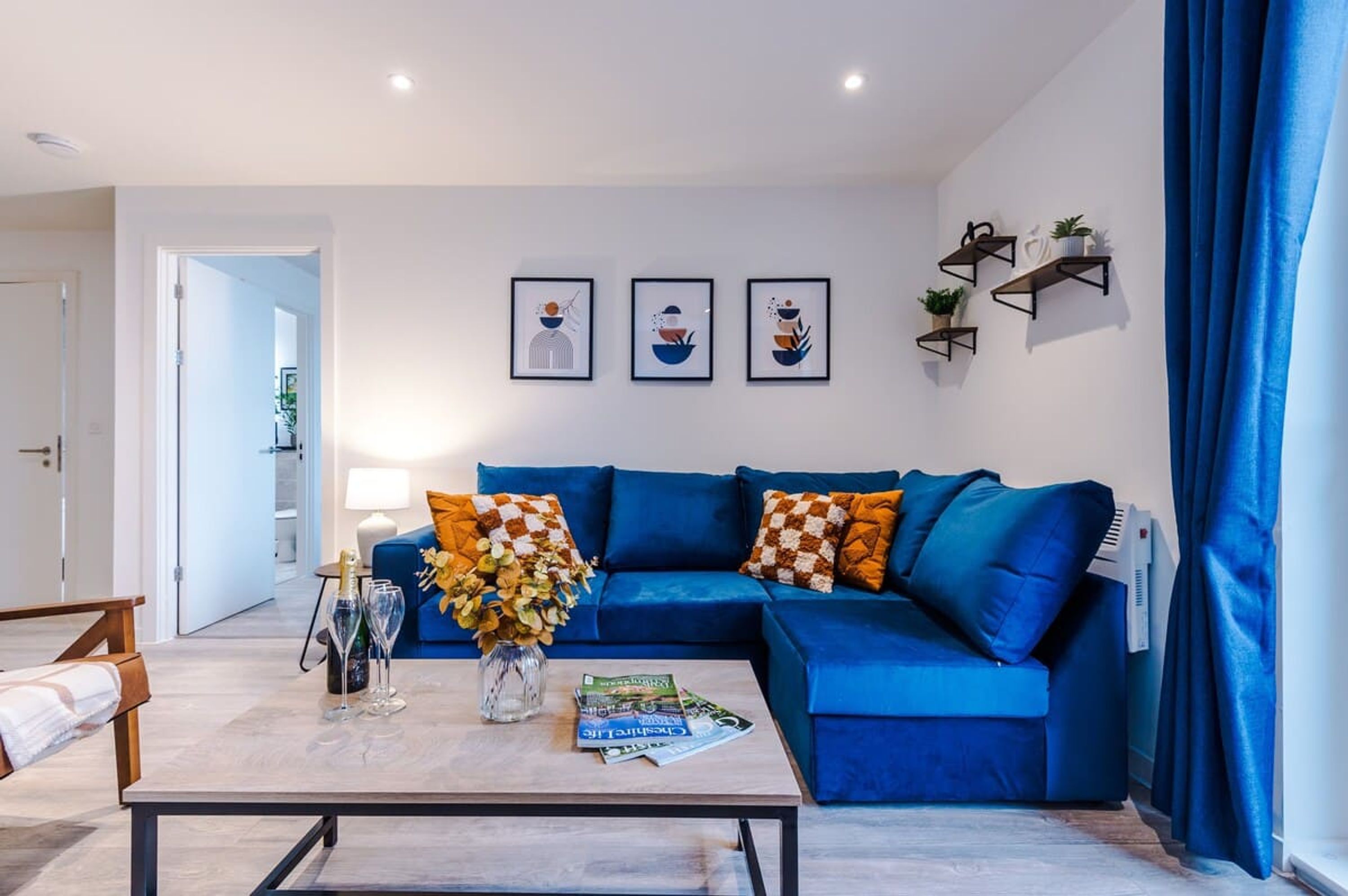 Ein Wohnzimmer mit einem blauen Sofa, einem Couchtisch mit Zeitschriften und einer Blumenvase, sowie drei gerahmten Kunstwerken an der Wand.
