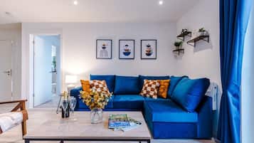 A living room with a blue sofa, a coffee table with magazines and a vase of flowers, and three framed artworks on the wall.