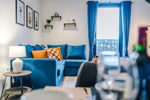 A modern living room with a blue sofa, a small round table with a lamp, and a window with blue curtains. - NEW Pristine 2-bed Flat Great Location Sleeps 6 (Liverpool)