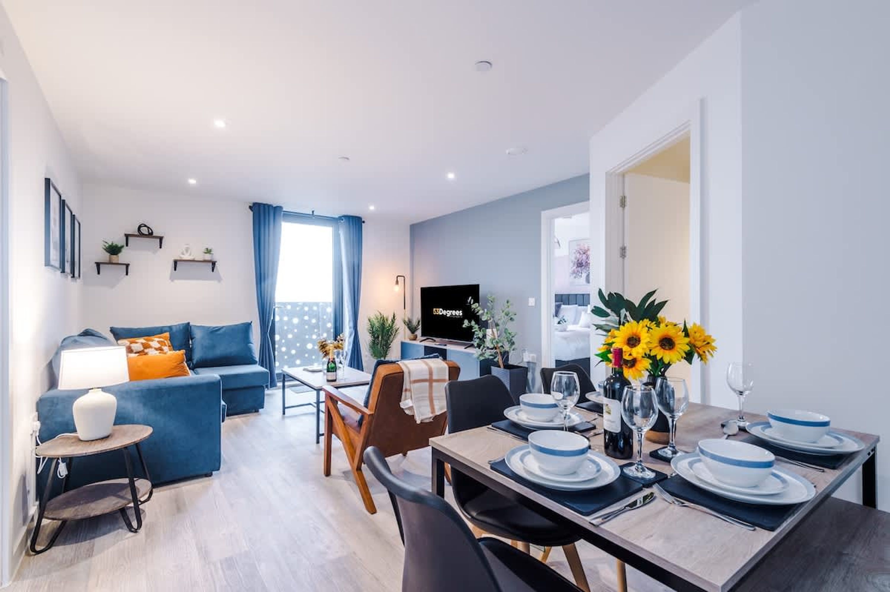 A modern living room with a sofa, dining table set for two, a TV, and a balcony with blue curtains.