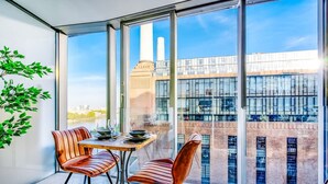 A modern dining area with a table and chairs, large windows, and a view of a building with a chimney. - NEW Pristine Apartment City Centre Amazing Views (London)