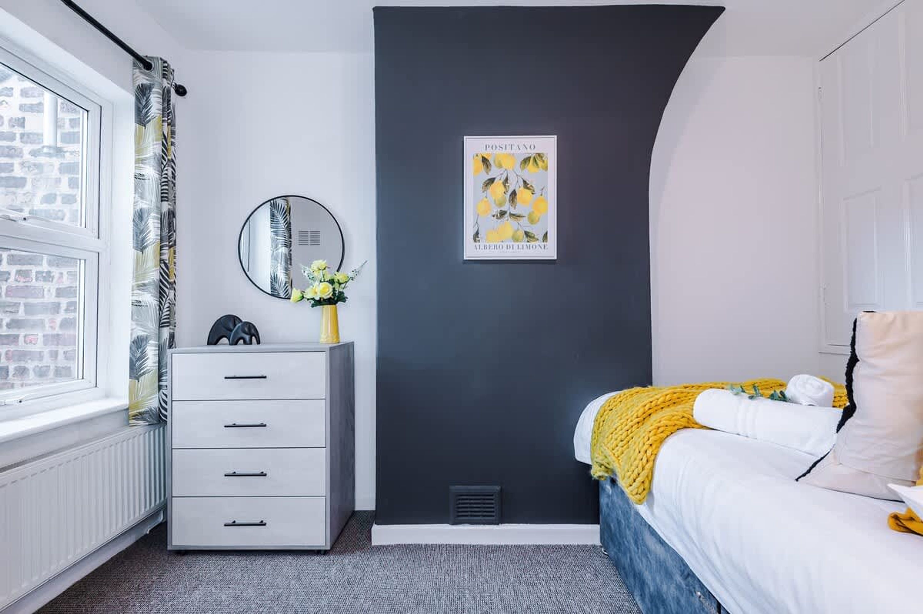 A bedroom with a bed, a dresser, a mirror, a framed picture, and a window with curtains.