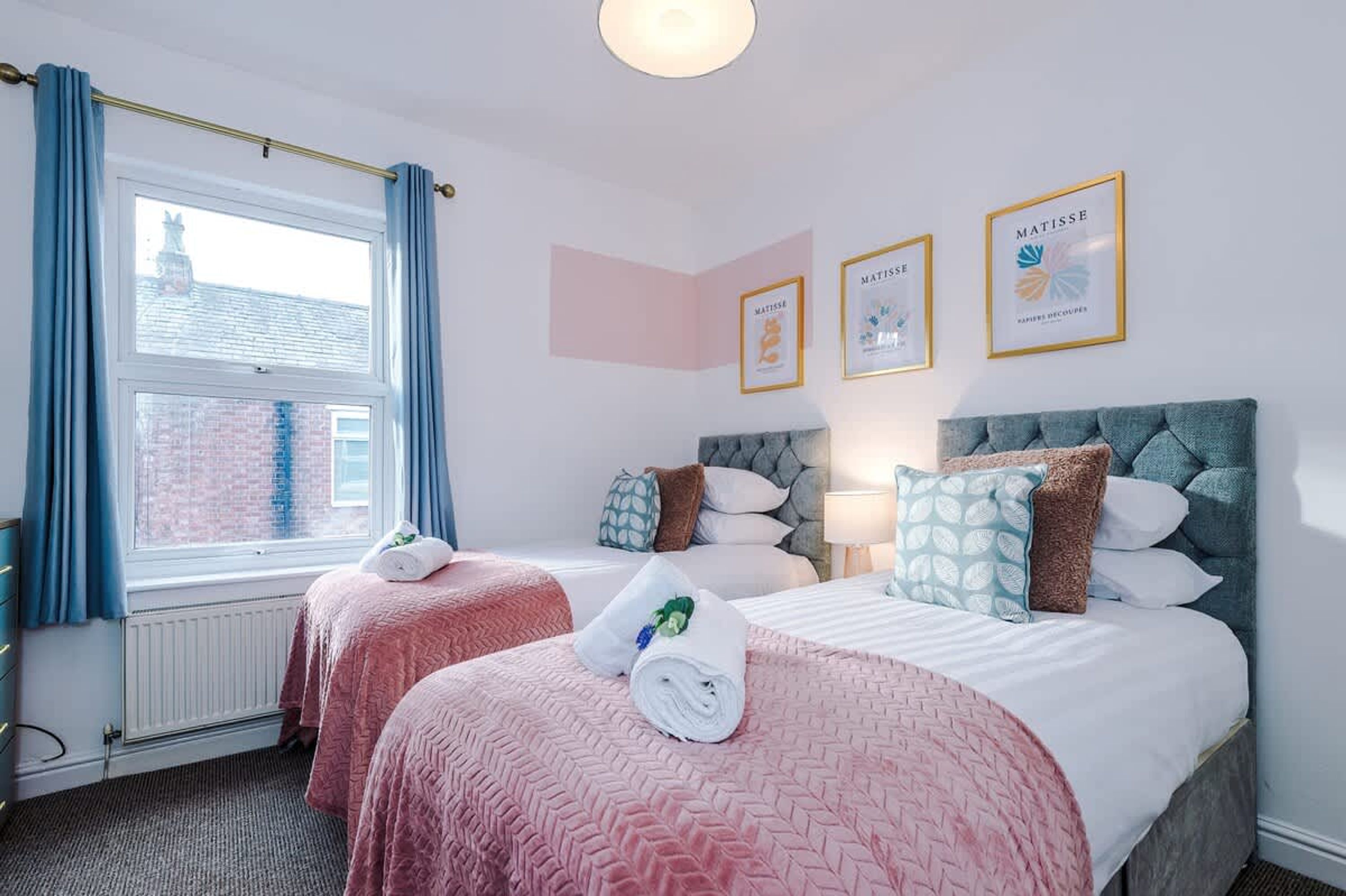 Une chambre d’hôtel avec deux lits, une fenêtre avec des rideaux et trois œuvres d’art encadrées accrochées au mur.