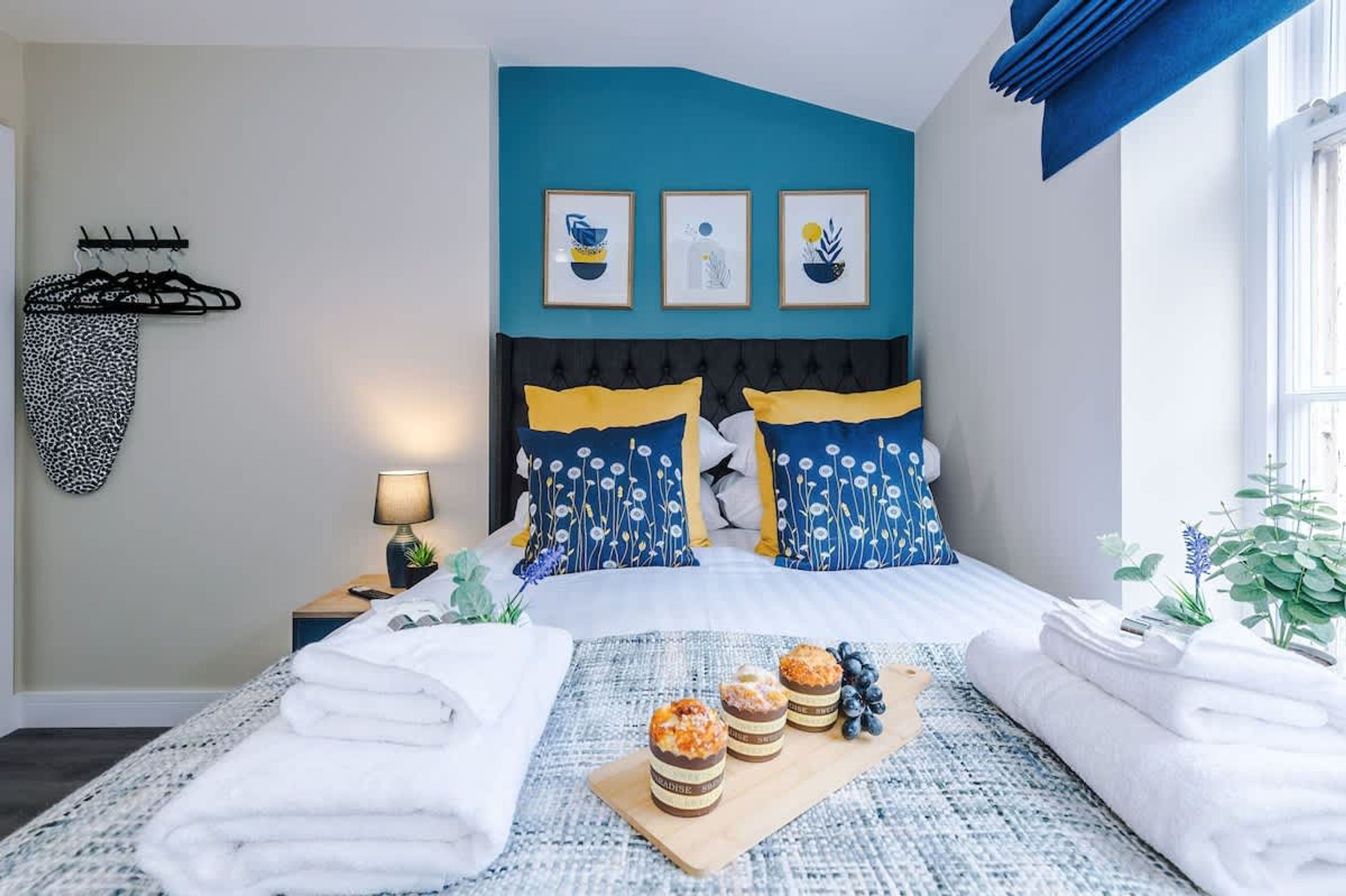 A bedroom with a bed, pillows, a nightstand, a lamp, a wall-mounted coat rack, and a window with curtains.