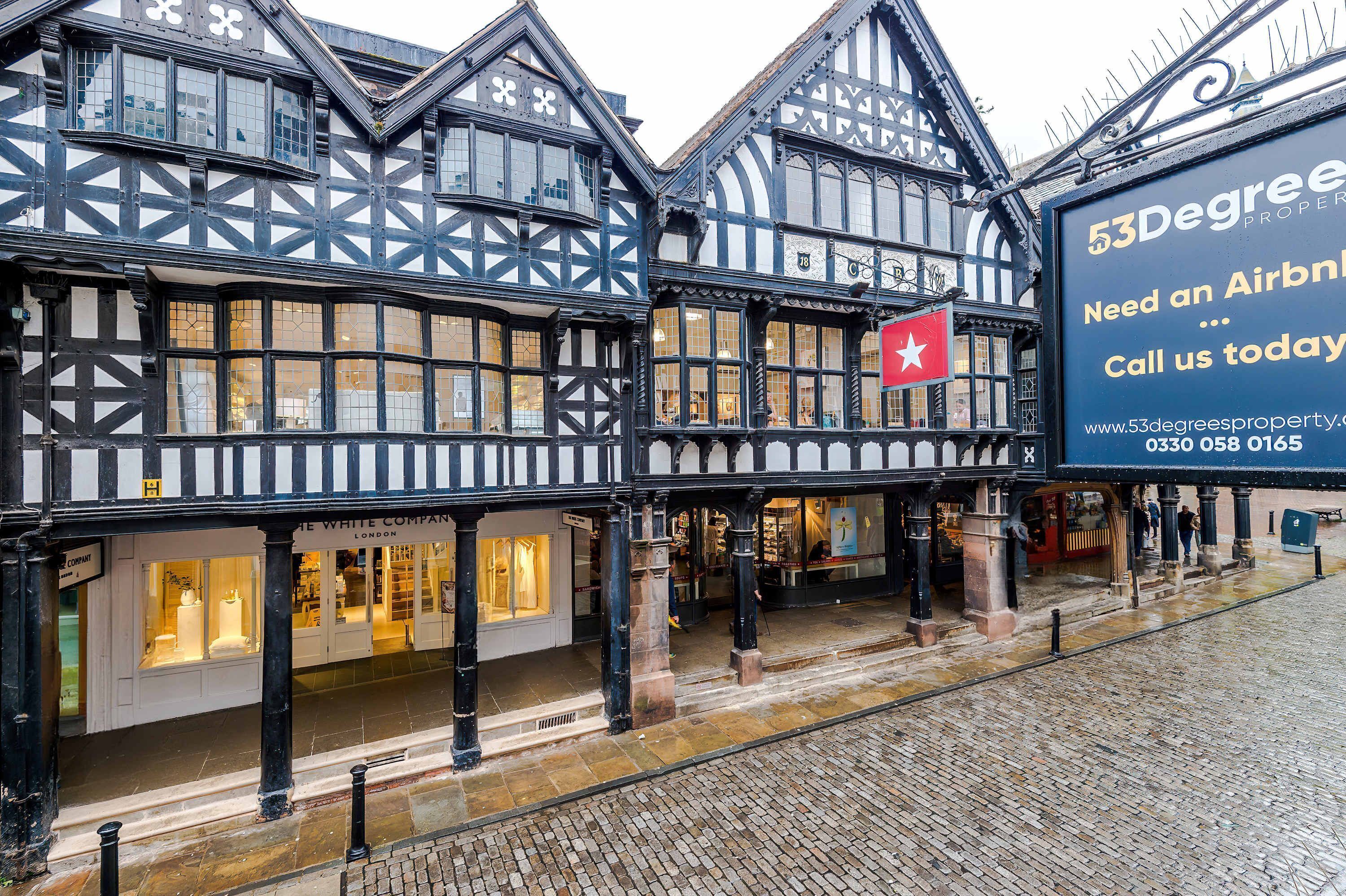 A traditional Tudor-style building with multiple ground-floor shops, a prominent red flag, and a large billboard advertising an Airbnb.
