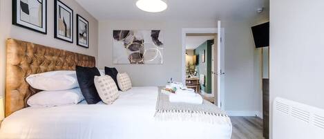 Une chambre à coucher avec une tête de lit capitonnée, du linge de lit blanc, une table de chevet, une lampe et un téléviseur à écran plat.