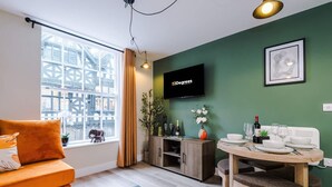 A modern living room with a sofa, a dining table set for four, a television, and a view of a cityscape through large windows. - NEW Stylish 1-bed Flat Great Location Sleeps 4 (Chester)