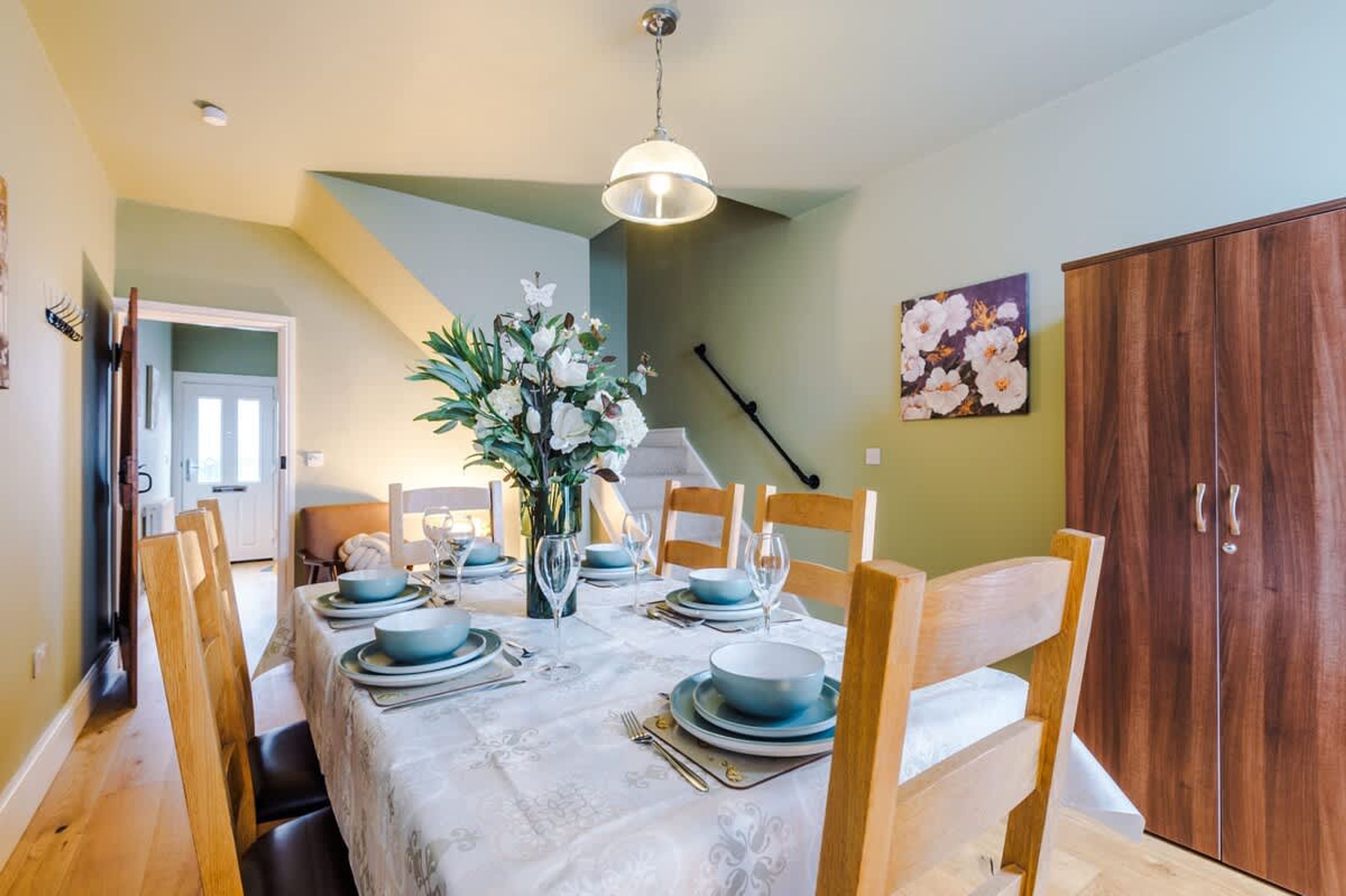 A dining room with a table set for four, wooden chairs, a vase with flowers, and a wooden cabinet.