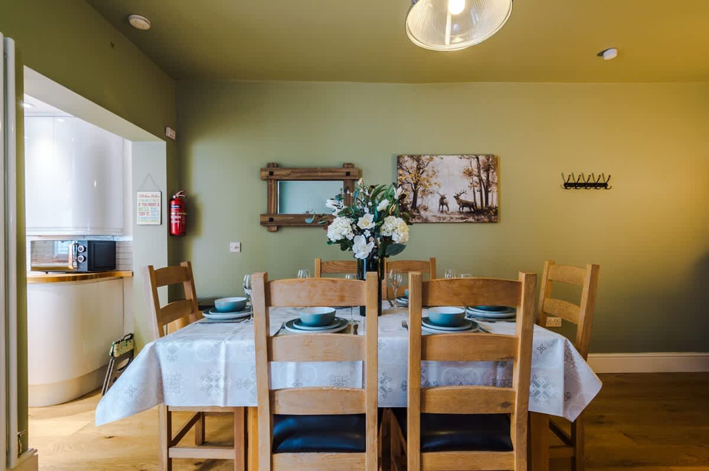 A dining area with a table set for four, wooden chairs, a vase with flowers, a fire extinguisher, and a painting on the wall.