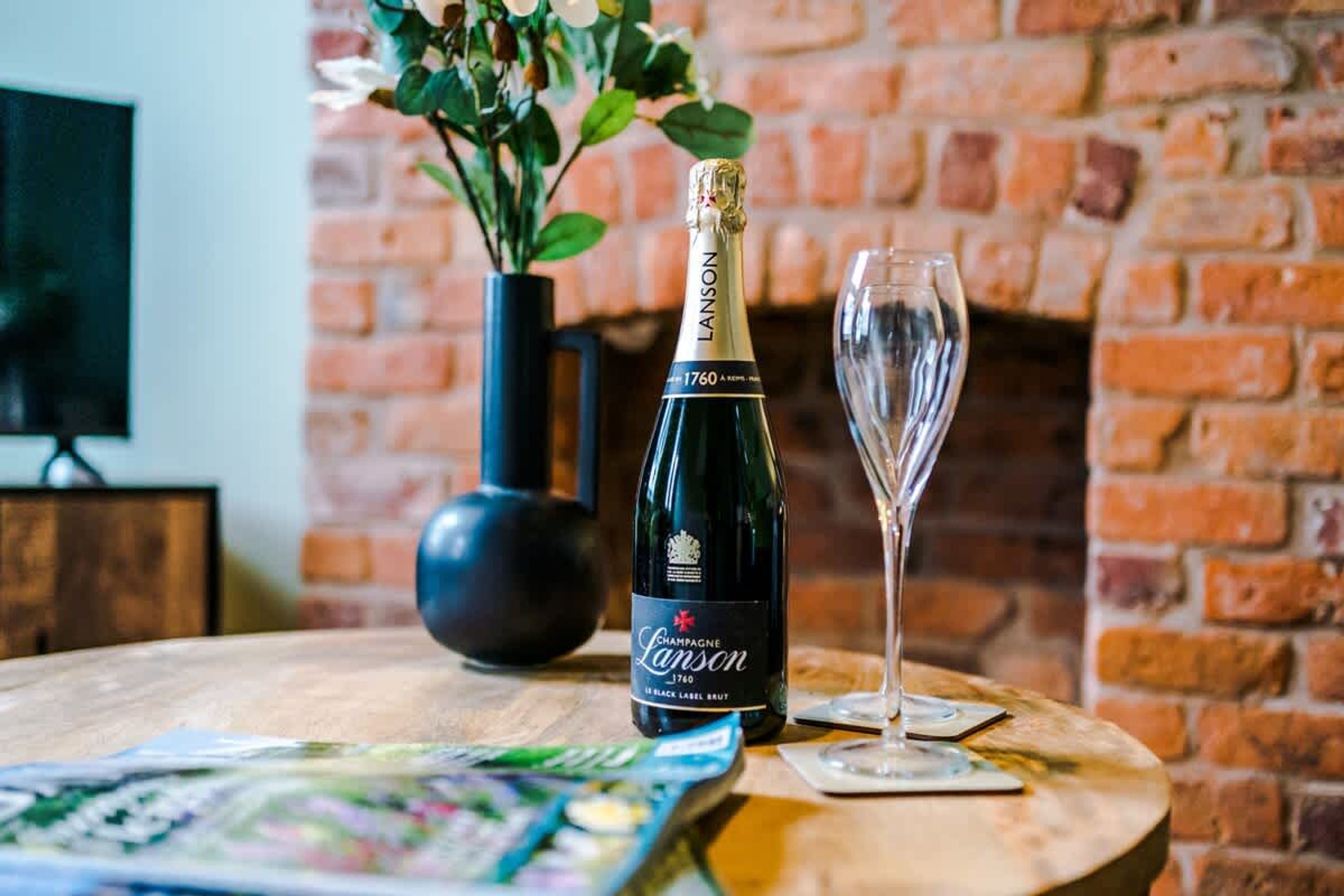 A bottle of champagne and a glass on a wooden table with a brick wall in the background.