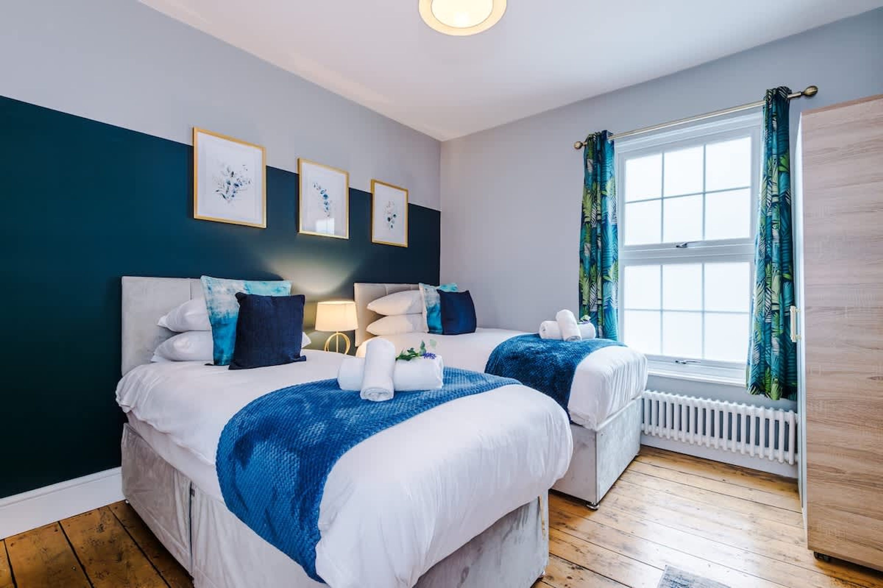 A hotel room with two beds, a window with curtains, a radiator, and framed artwork on the wall.