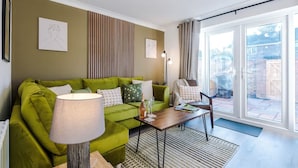 Ett modernt vardagsrum med en grön soffa, ett soffbord och en lampa. På väggen hänger två inramade konstverk, och en skjutdörr i glas leder ut till ett utomhusområde.