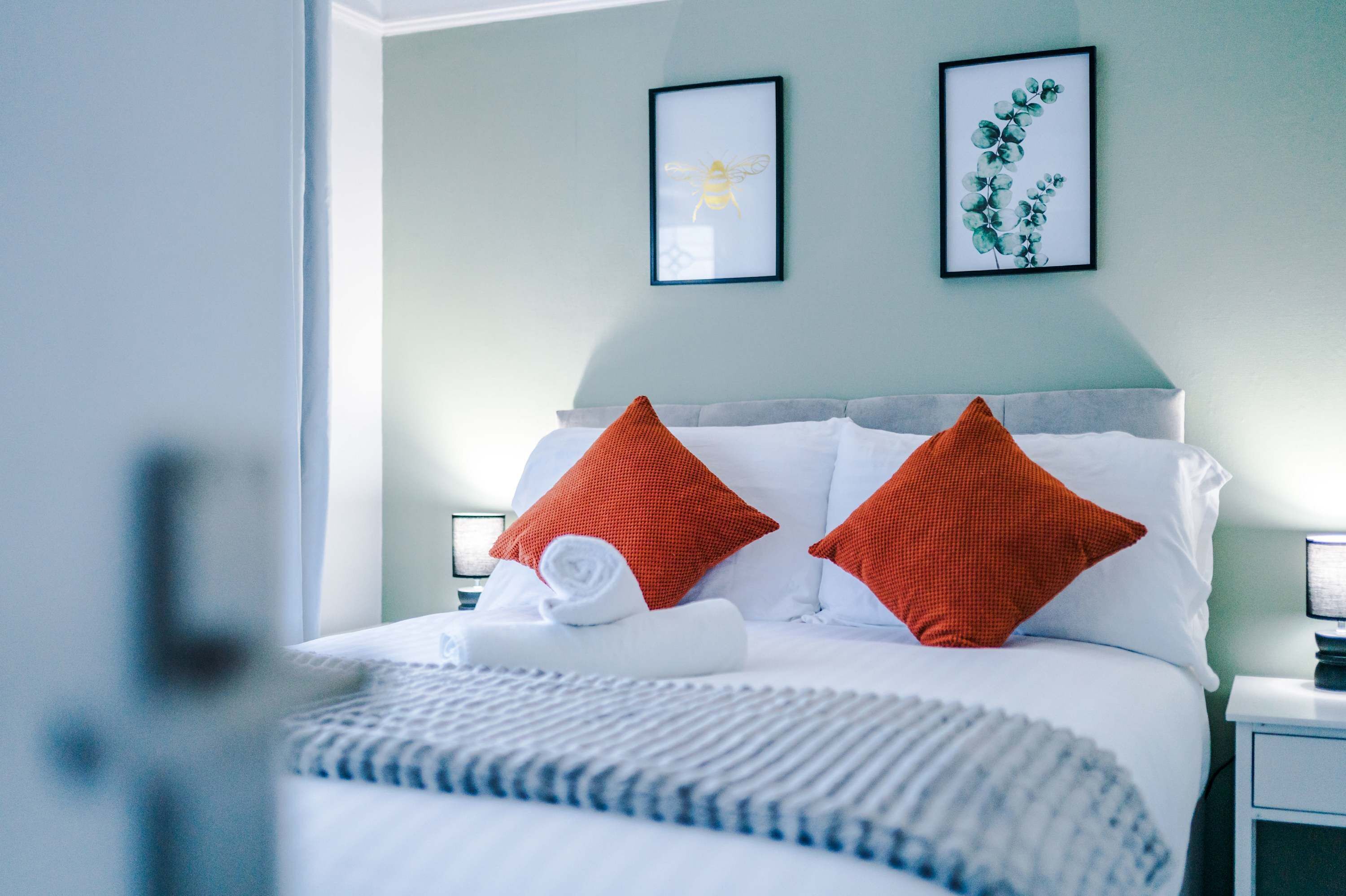 A neatly made bed with white and red pillows, a rolled white towel, and a blue and white striped blanket. There are two framed pictures on the wall above the bed.