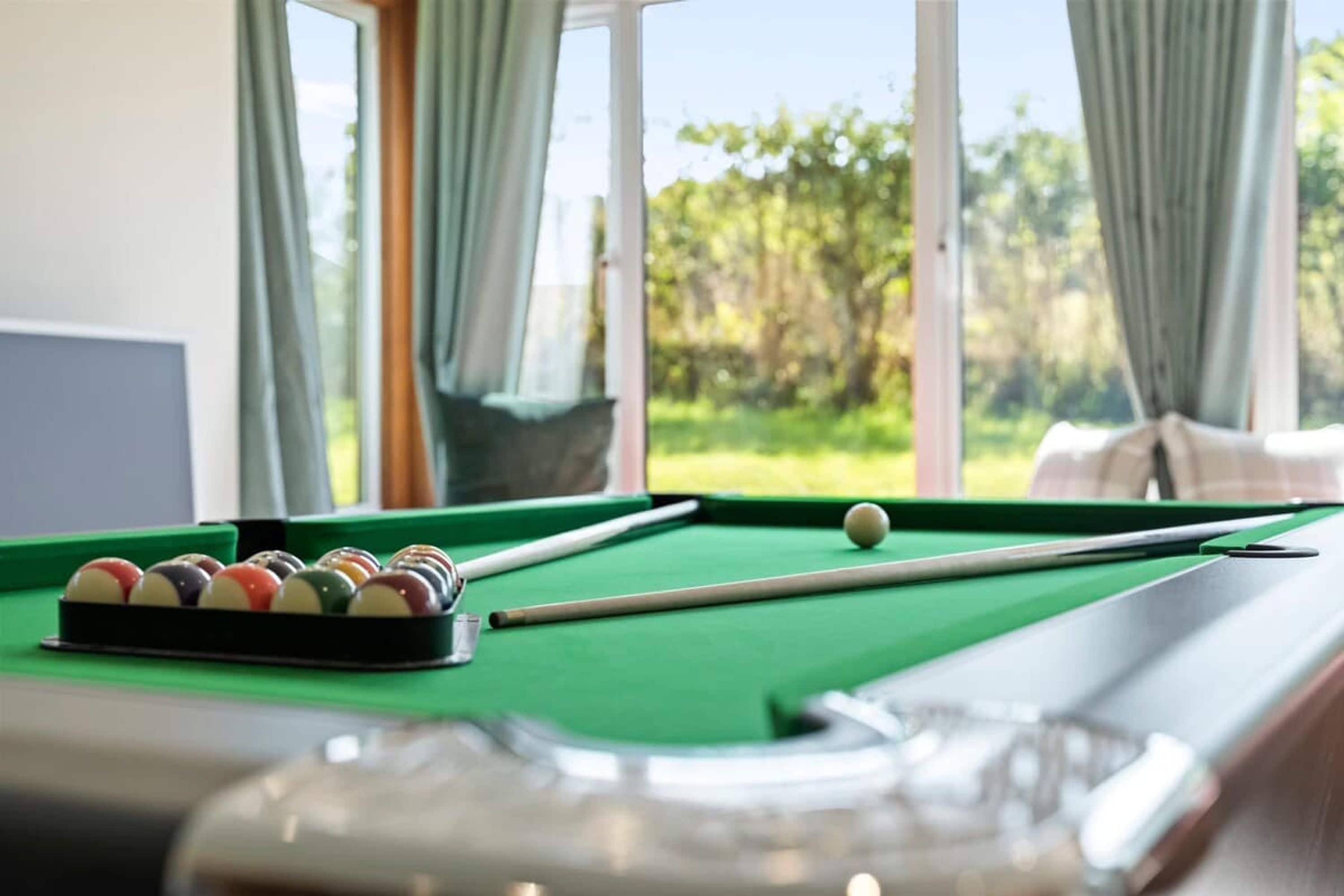 A pool table with balls and a cue stick, a window with curtains, and a view of greenery outside.