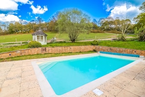 Une piscine rectangulaire avec un mur en pierre et un petit kiosque situé dans un espace verdoyant.