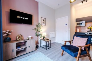 Een moderne woonkamer met een flatscreen televisie, een blauwe fluweelstoel, een houten console, een klein tafeltje met een lamp en een potplant.