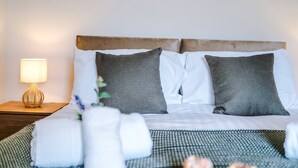 Een net opgemaakt bed met witte en grijze kussens, een bedovertrek met een groen-wit patroon en een klein tafeltje met een lamp ernaast.