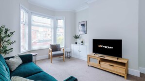 Een moderne woonkamer met een blauwgroene bank, een grijze fauteuil, een televisie op een houten dressoir, een kast en een raam met jaloezieën.
