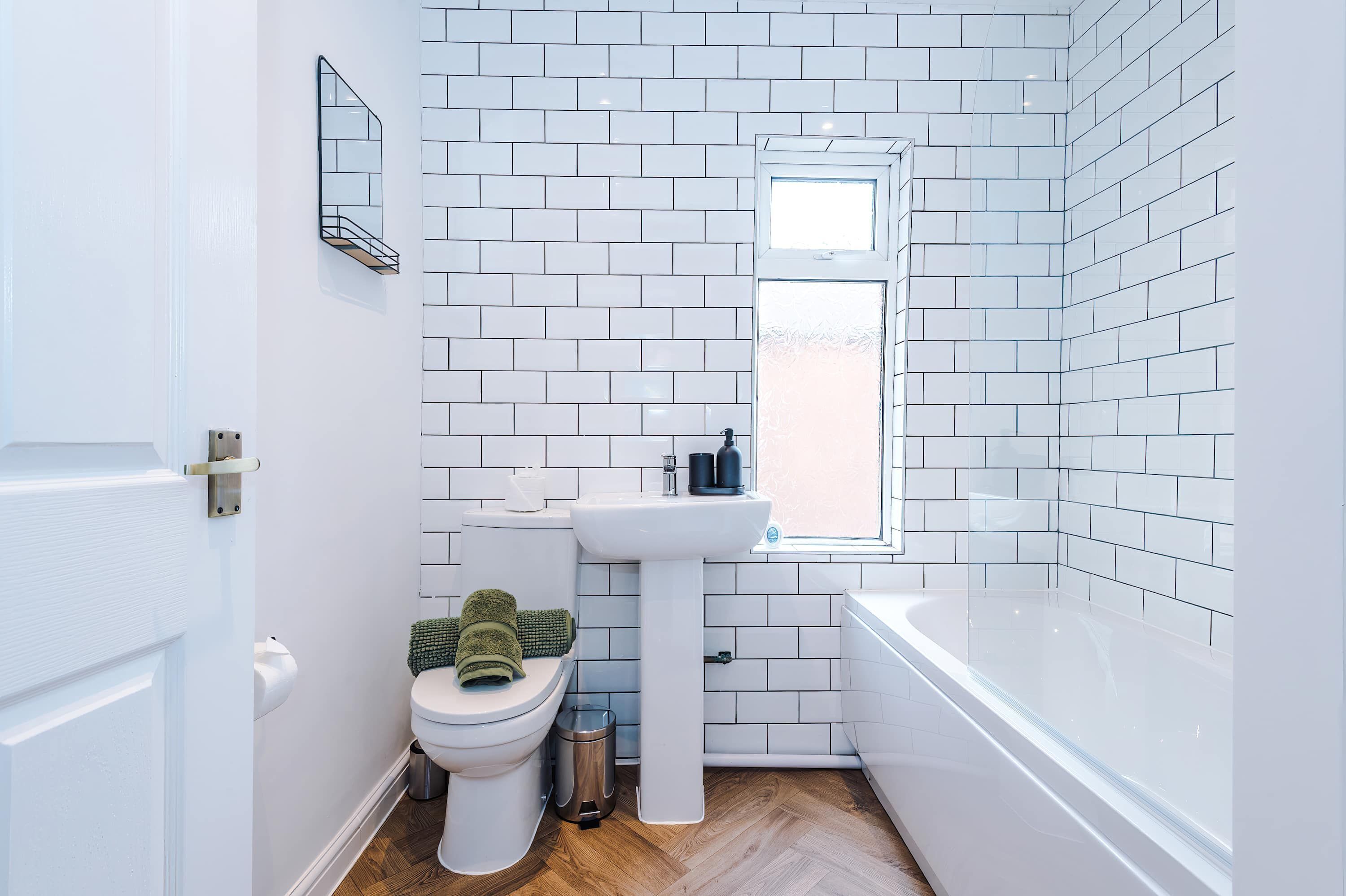 A bathroom with white tiled walls, a white sink, a toilet, a window, and a bathtub.