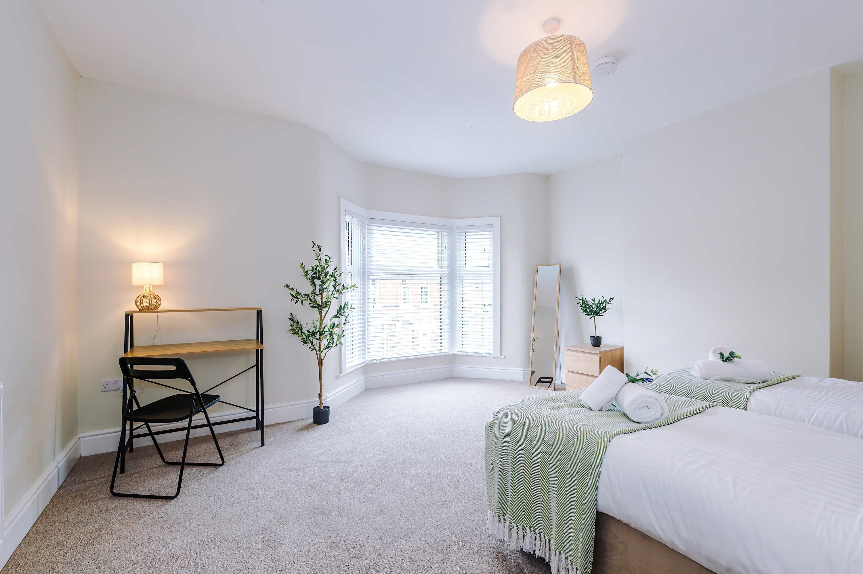 A bedroom with a desk, two beds, a window, a hanging light fixture, and a potted plant.
