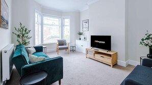 Een moderne woonkamer met een blauwgroene bank, een houten tv-meubel, een klein bijzettafeltje en een radiator.