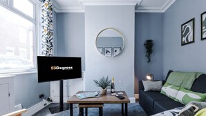 Un salon comprenant un téléviseur, un miroir rond, un canapé et une table basse ornée d’une plante et d’une bouteille.