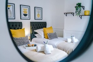 A hotel room with two beds, a mirror, artwork on the wall, and a small shelf with a plant. - NEW Spacious Home Ideal for Groups Free Parking (Newcastle-Under-Lyme)
