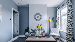 Un coin repas avec une table dressée pour deux personnes, une horloge murale et une fenêtre ornée de rideaux à motifs.