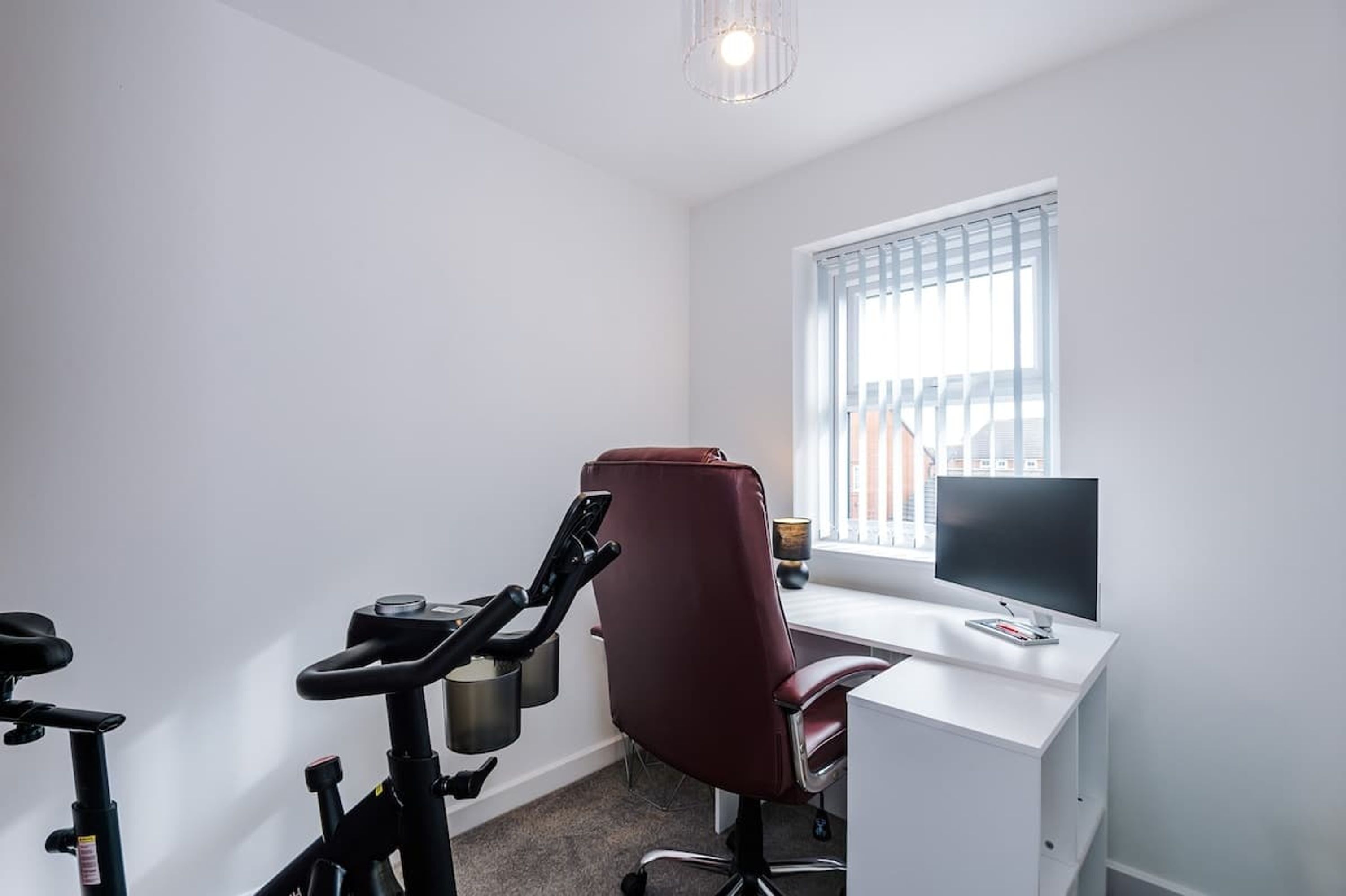 A room with a stationary bike, a desk with a computer, and a window with blinds.