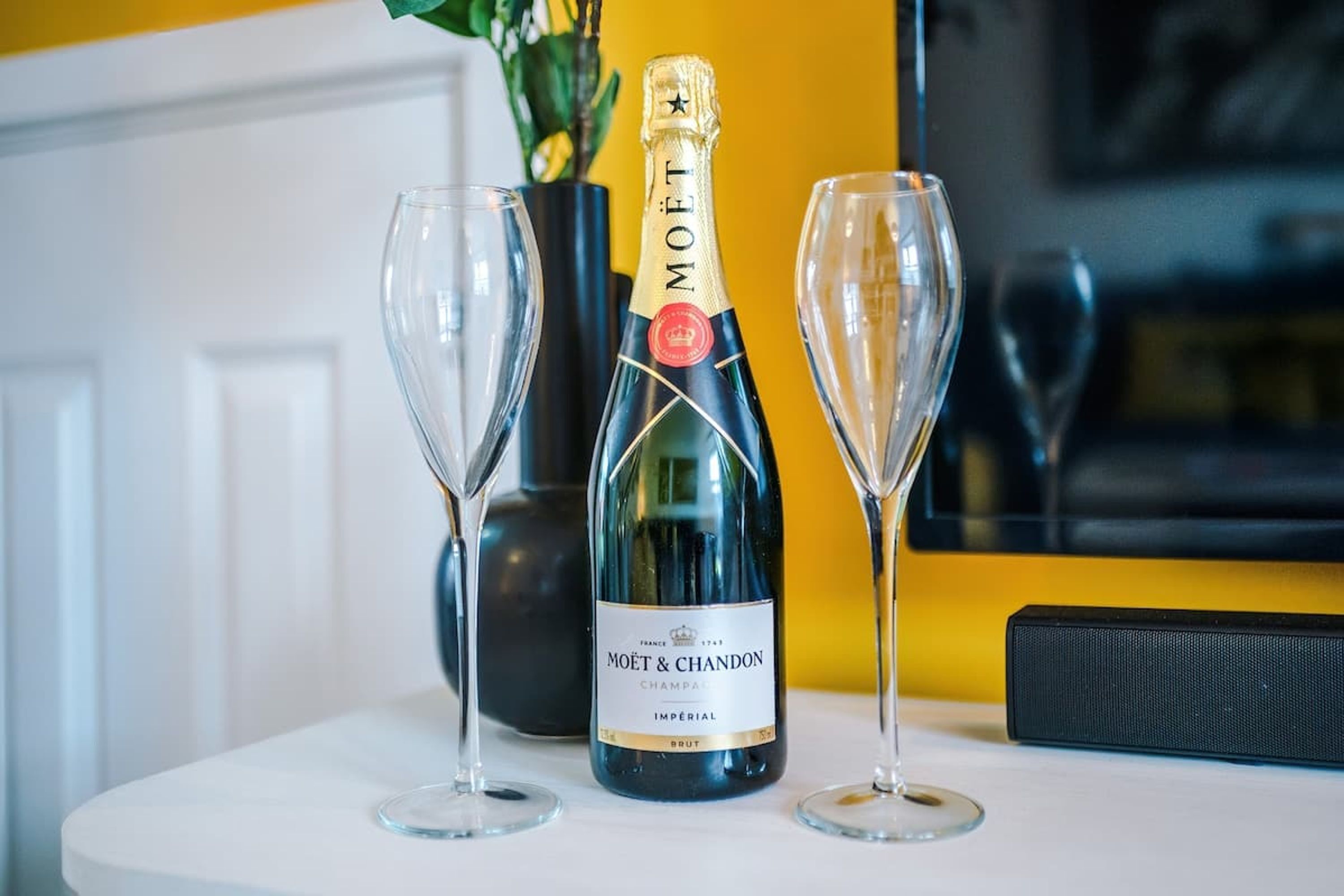 A bottle of Moët & Chandon champagne, two empty wine glasses, and a black speaker on a white table.